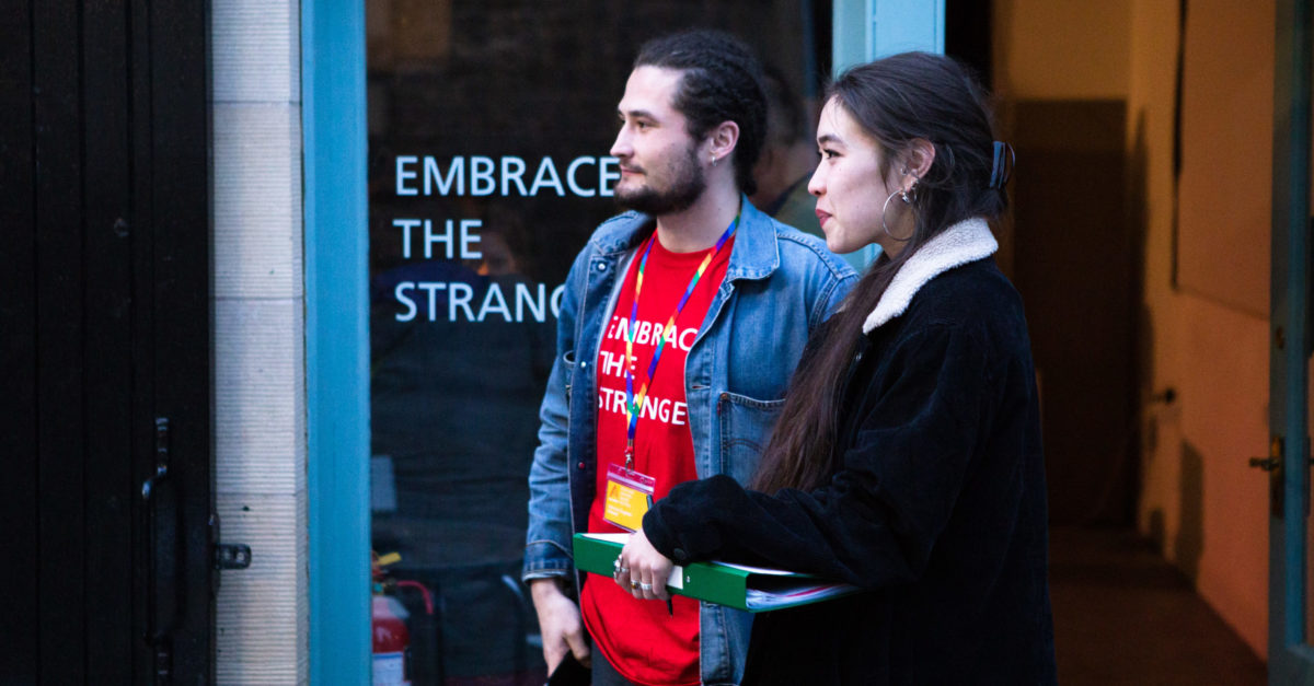 Two people in their 20s stand at the entrance of a venue at Alchemy Film and Moving Image Festival ready to greet and welcome attendees. One wears an Alchemy t-shirt that reads "Embrace the Strange" and an LGBTQ pride lanyard. The other holds a clipboard. The words "Embrace the Strange" are also printed on the window of the festival venue.